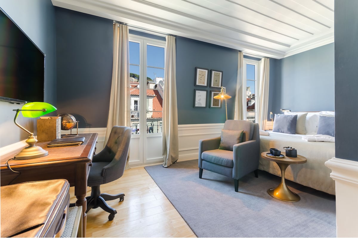 Una habitación de hotel moderna con un escritorio de madera, un sillón cómodo, una pequeña mesa redonda con lámpara y una cama con vistas a la ciudad.