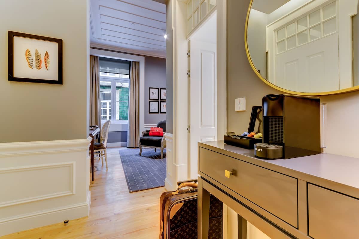 Habitación de hotel con piso de madera, un espejo grande, un escritorio y una silla. Hay una puerta que conduce a otra habitación y una ventana con cortinas.