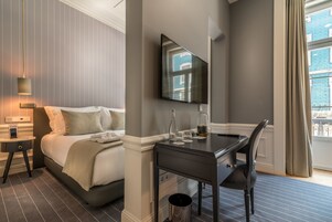 A modern hotel room with a bed, bedside table, desk, chair, and a large window with curtains.