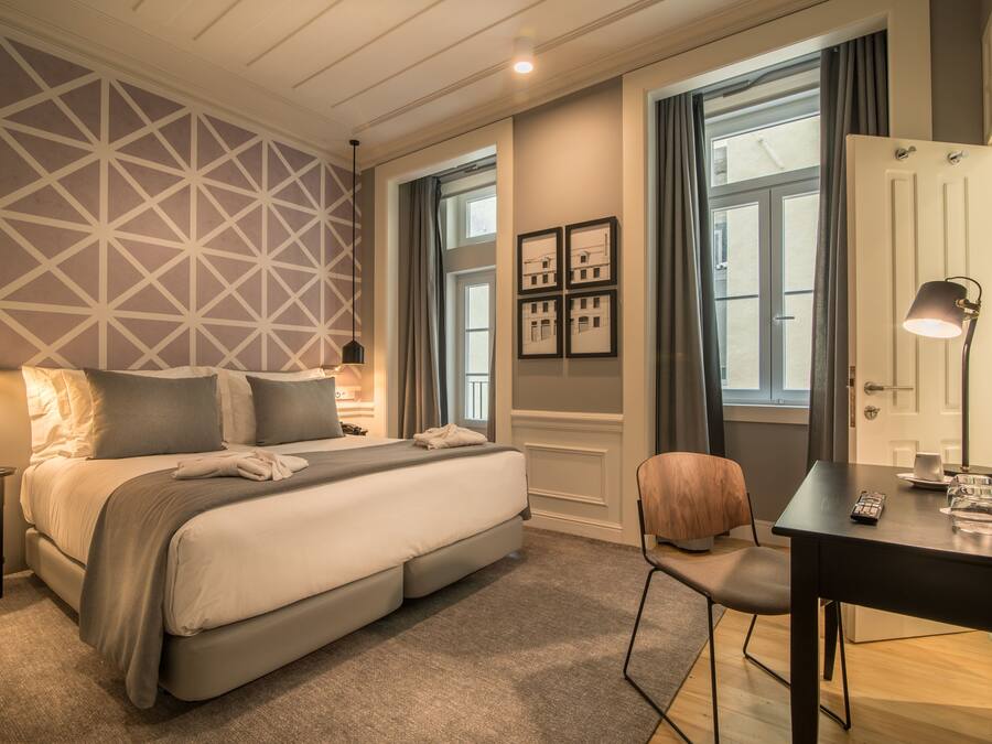 Una habitación de hotel moderna con una cama grande, un escritorio con lámpara y una silla. Hay cuadros enmarcados en la pared y una ventana con cortinas.