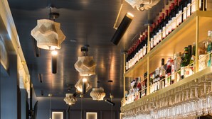 Eine Bar mit Theke, Barhocker, hängenden Lampen und Regalen voller Flaschen und Gläsern.