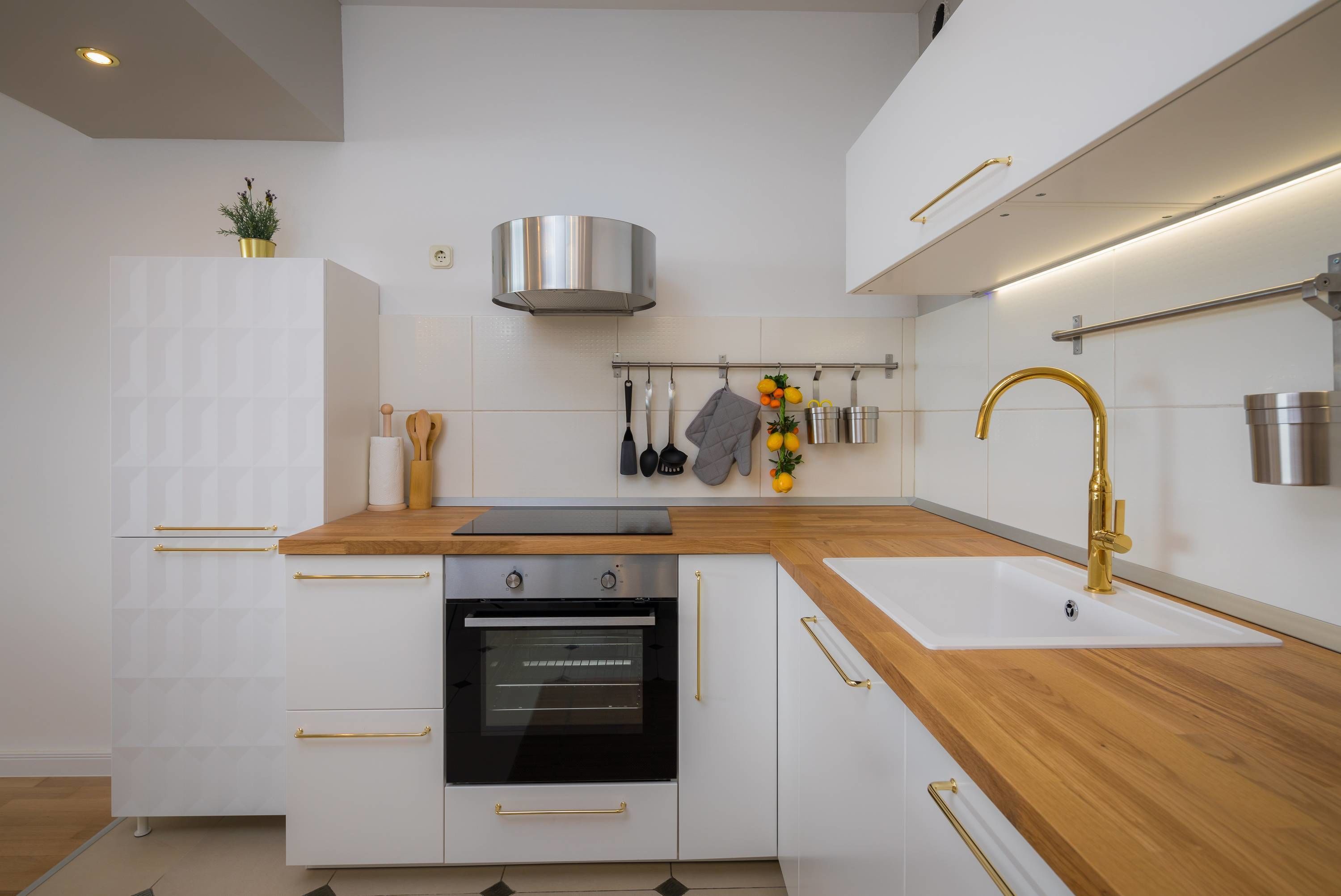 A modern kitchen with white cabinetry, a wooden countertop, a stainless steel range hood, and a gold faucet.