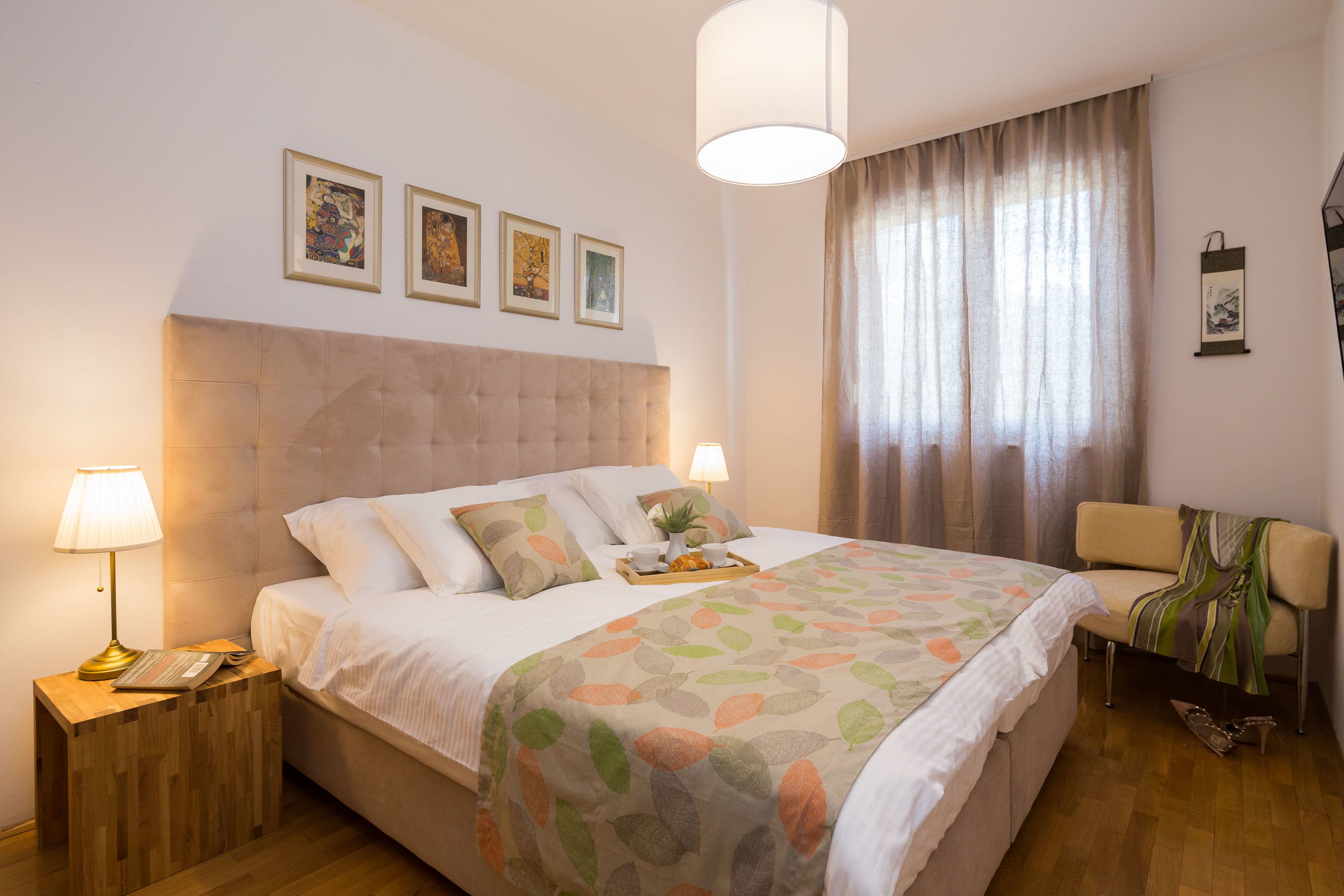 A neatly arranged hotel room with a large bed, bedside lamps, a small table with a tray, and a chair with a blanket.