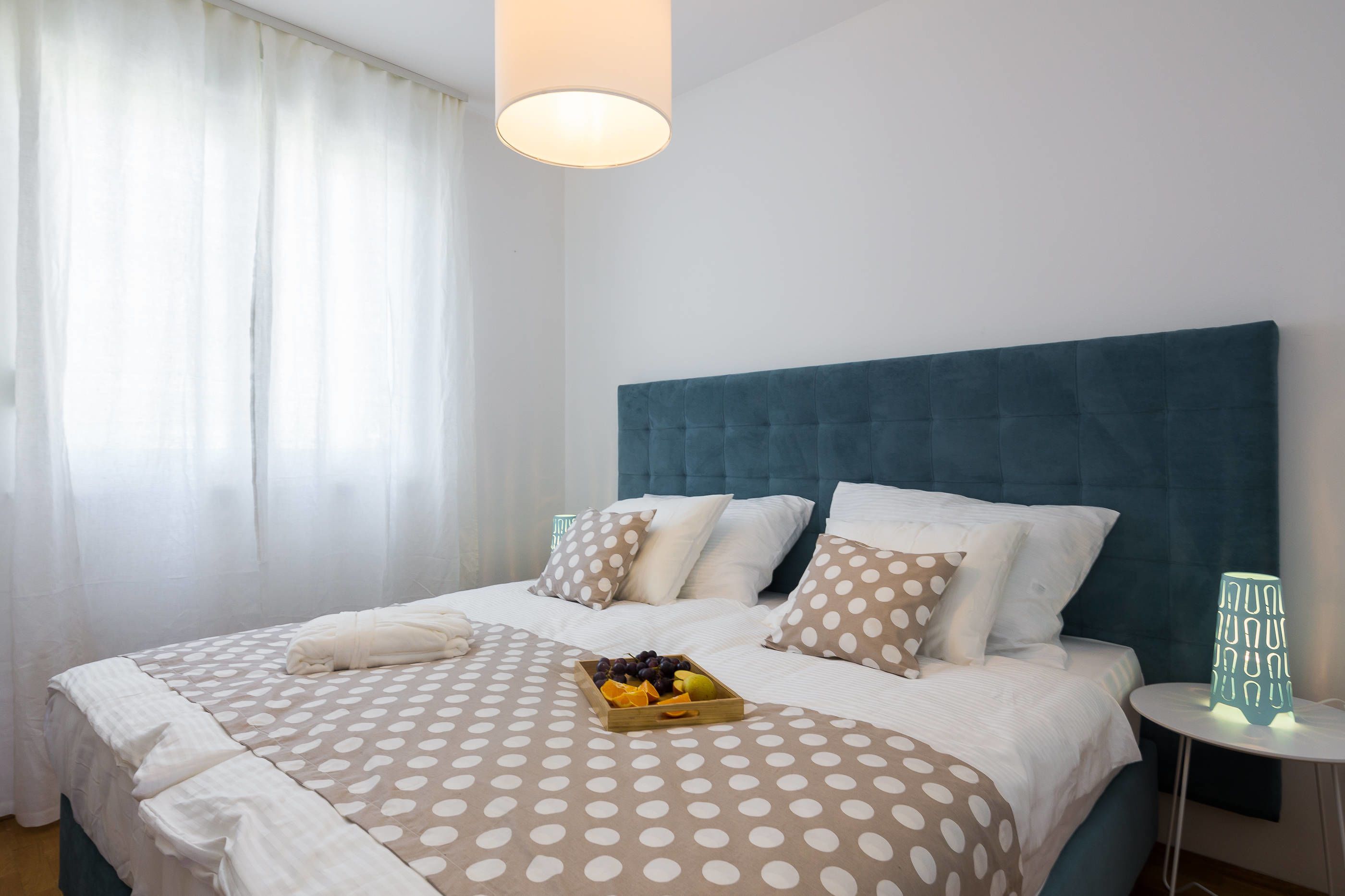 Un lit bien fait, recouvert d’une couvre-lit à pois, avec un plateau de fruits et une lampe de chevet.