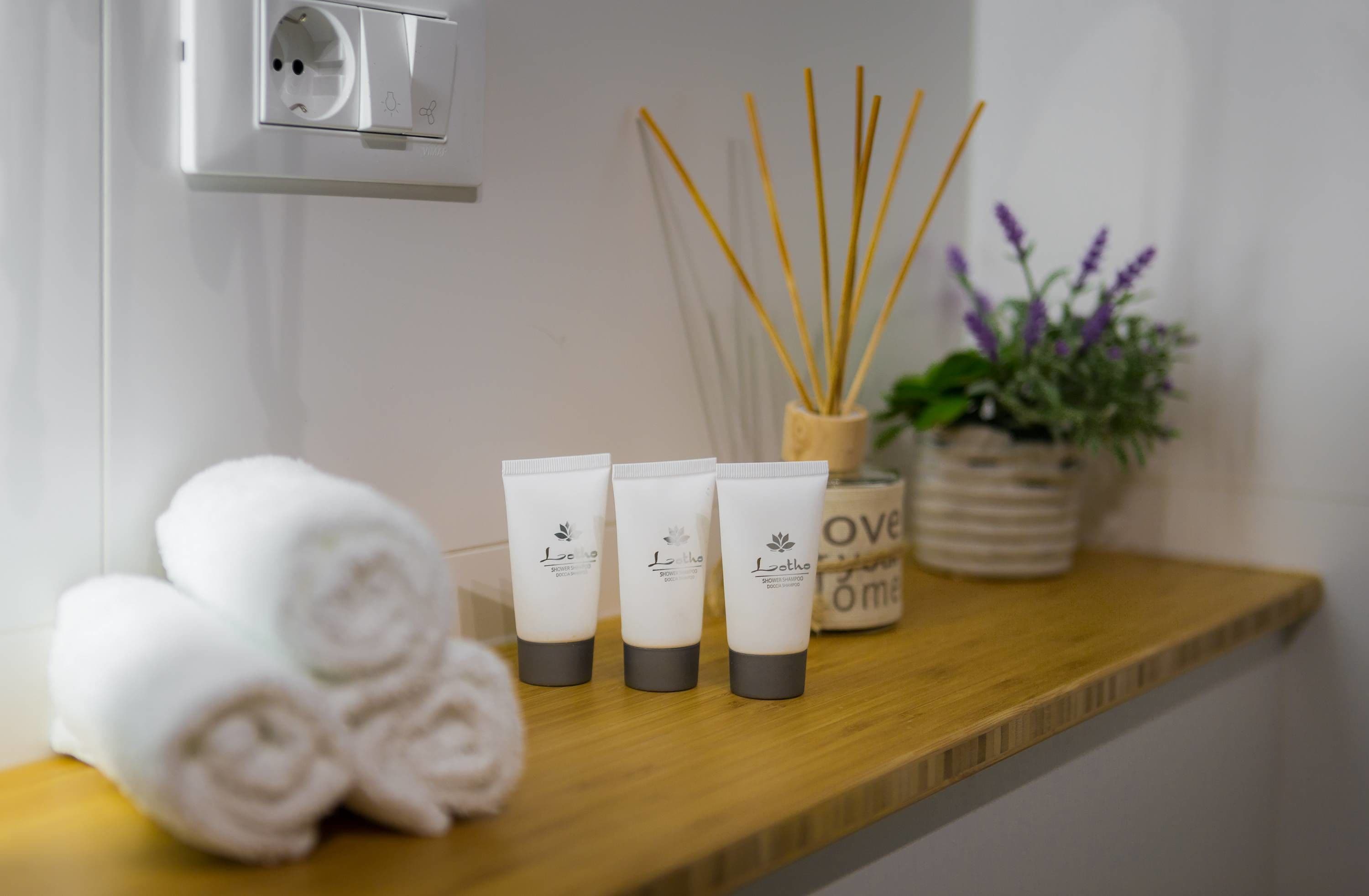 Un estante de baño con toallas enrolladas, tres tubos de loción, difusores de aroma y una planta en maceta.