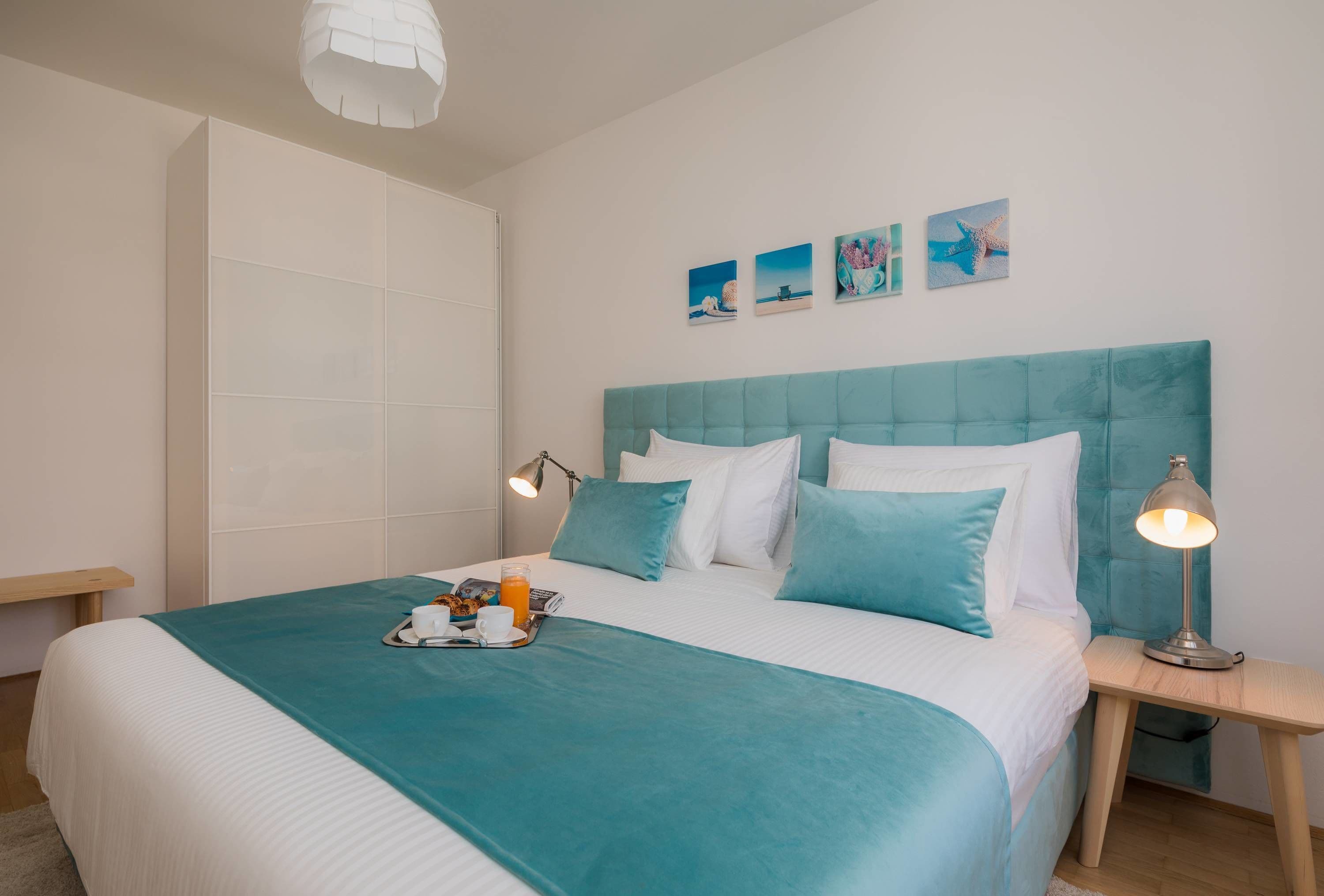 A neatly made bed with a teal bedspread, white pillows, and a tray with a cup and orange juice.