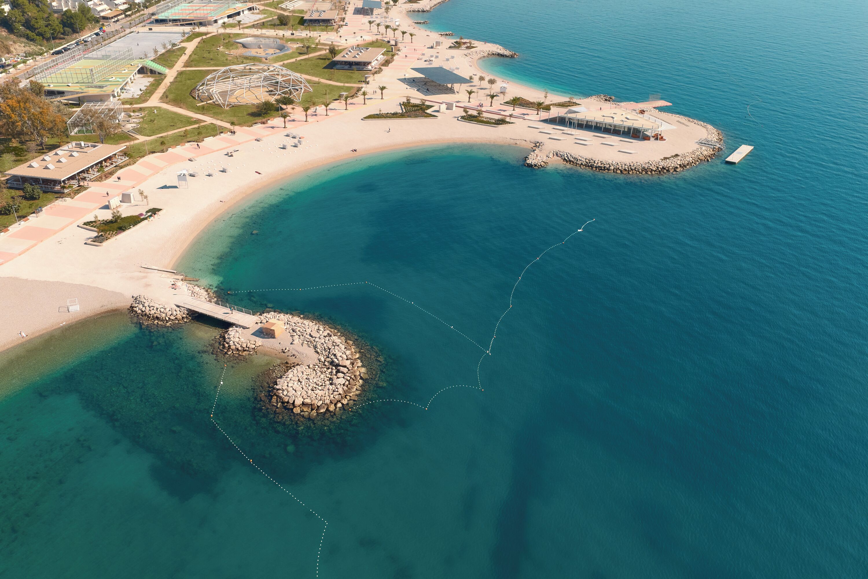 An aerial view of a coastal area with a sandy beach, clear blue waters, and a rocky outcrop.