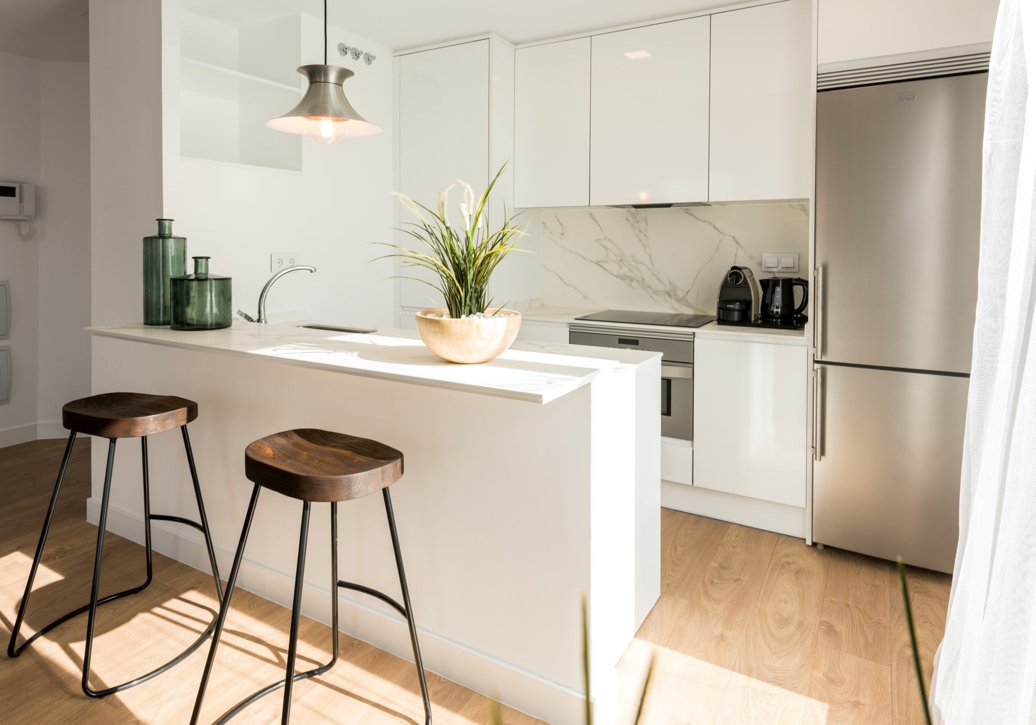 A modern kitchen with white cabinetry, a marble backsplash, a stainless steel refrigerator, and two wooden stools.
