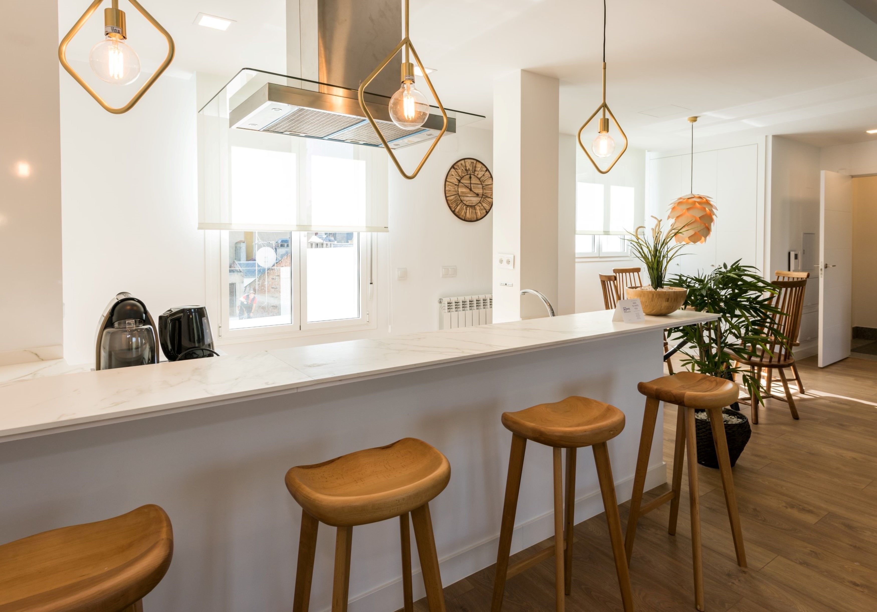 A modern kitchen with a white countertop, wooden stools, pendant lighting, and a view of the outside.