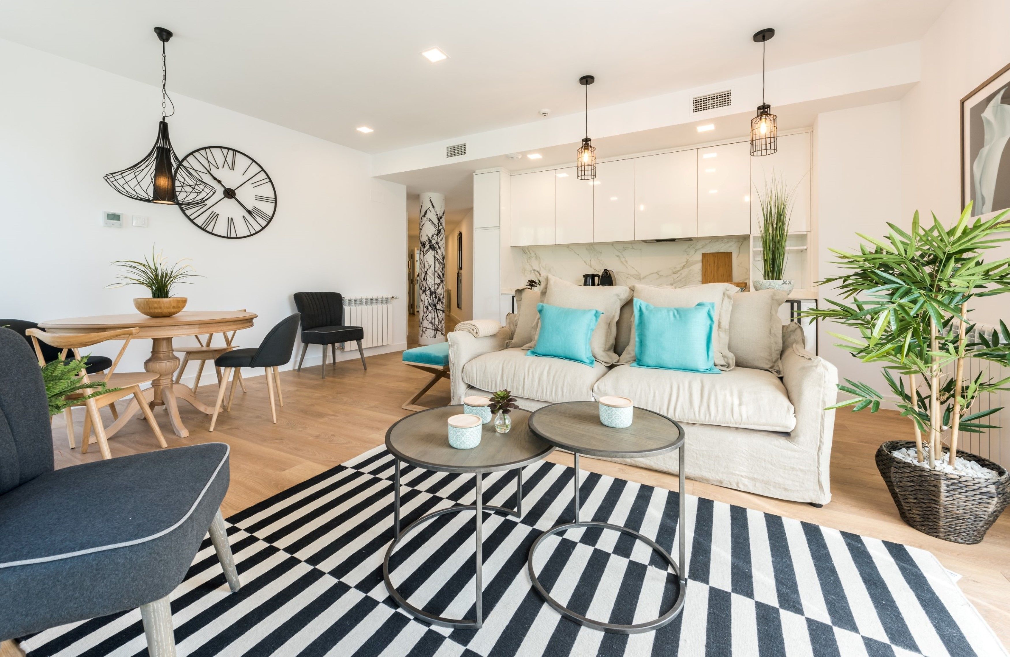 Una sala de estar moderna con sofá, mesa de centro y sillas. Hay un comedor con mesa y sillas. La habitación tiene un reloj en la pared y lámparas colgantes.