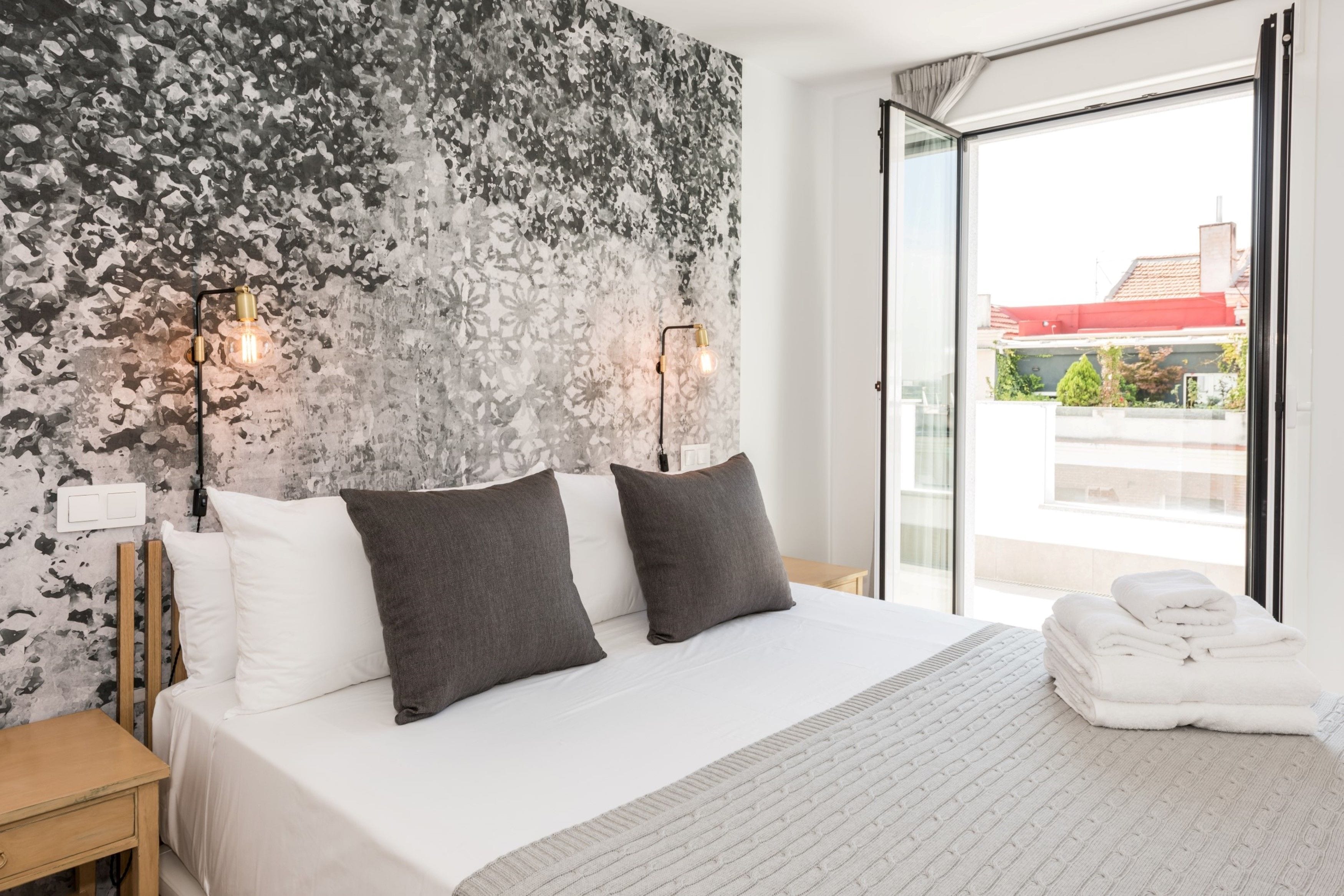 Una habitación de hotel moderna con una cama grande, dos almohadas, una mesita de noche y vistas al exterior a través de un ventanal.