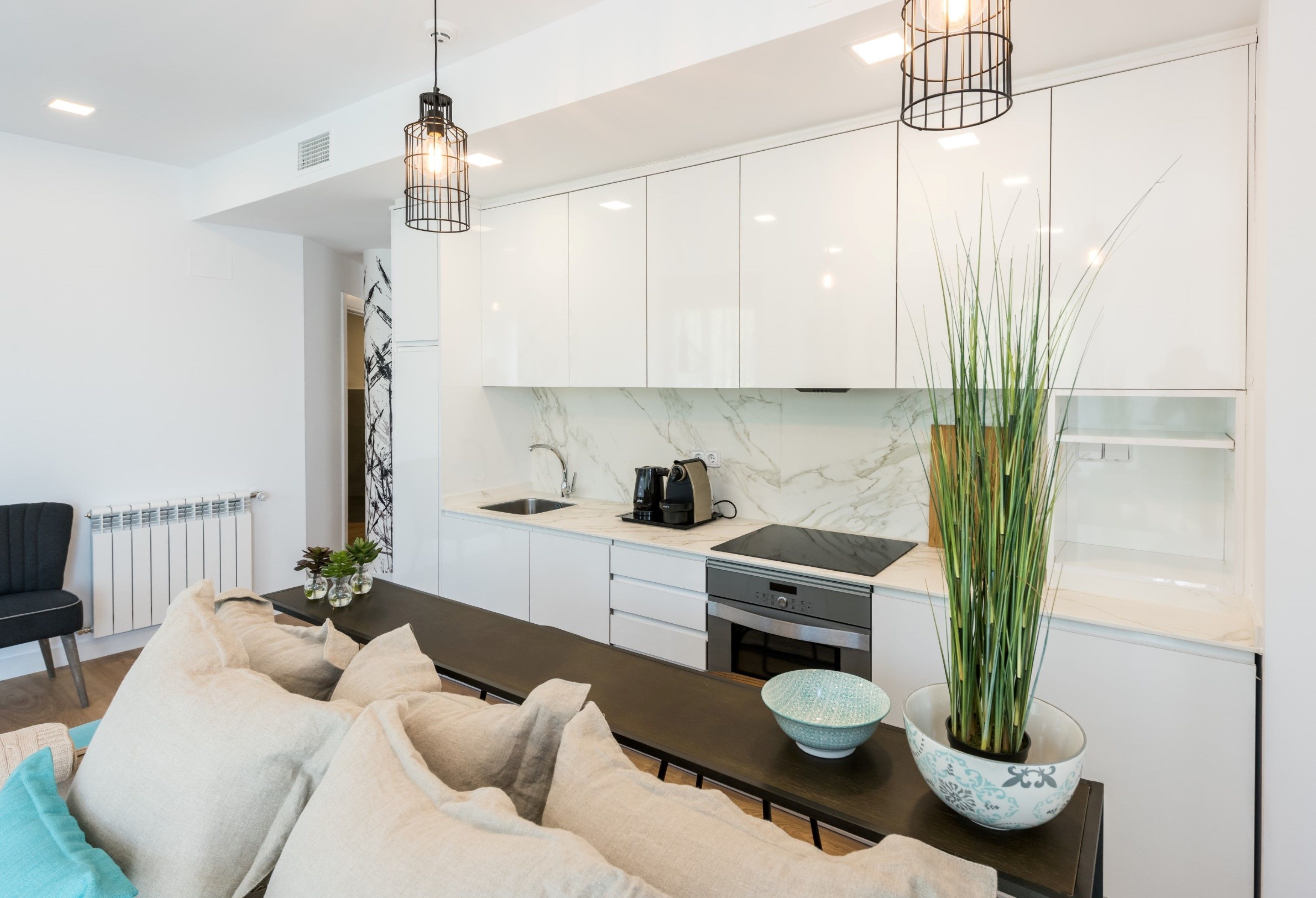 A modern kitchen with white cabinets, a marble countertop, and a black stove. There is a dining area with a dark wooden table and beige cushions.