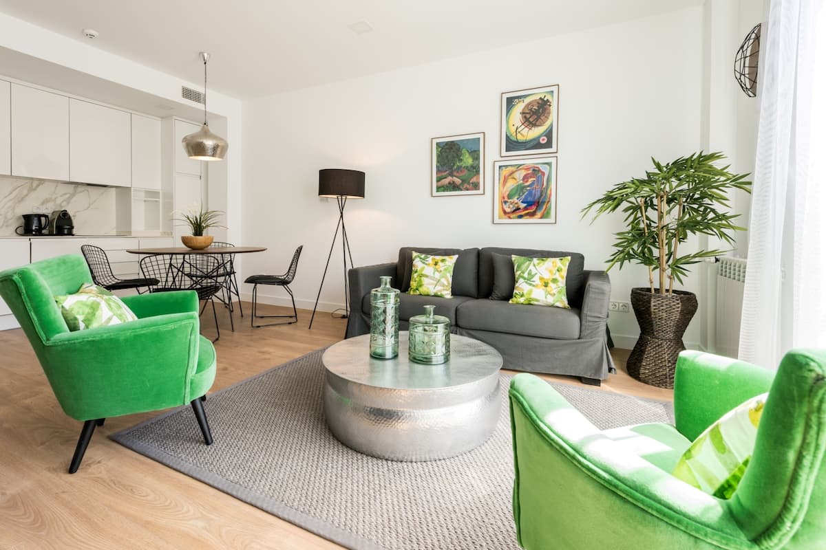 Una sala de estar moderna con un sofá gris, sillones verdes y una mesa de centro redonda. Hay una zona de comedor con una mesa y sillas, y una cocina al fondo.