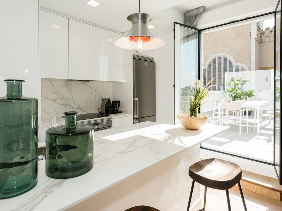 Una cocina moderna con encimeras de mármol blanco, un refrigerador de acero inoxidable y un amplio ventanal con vistas a un patio.