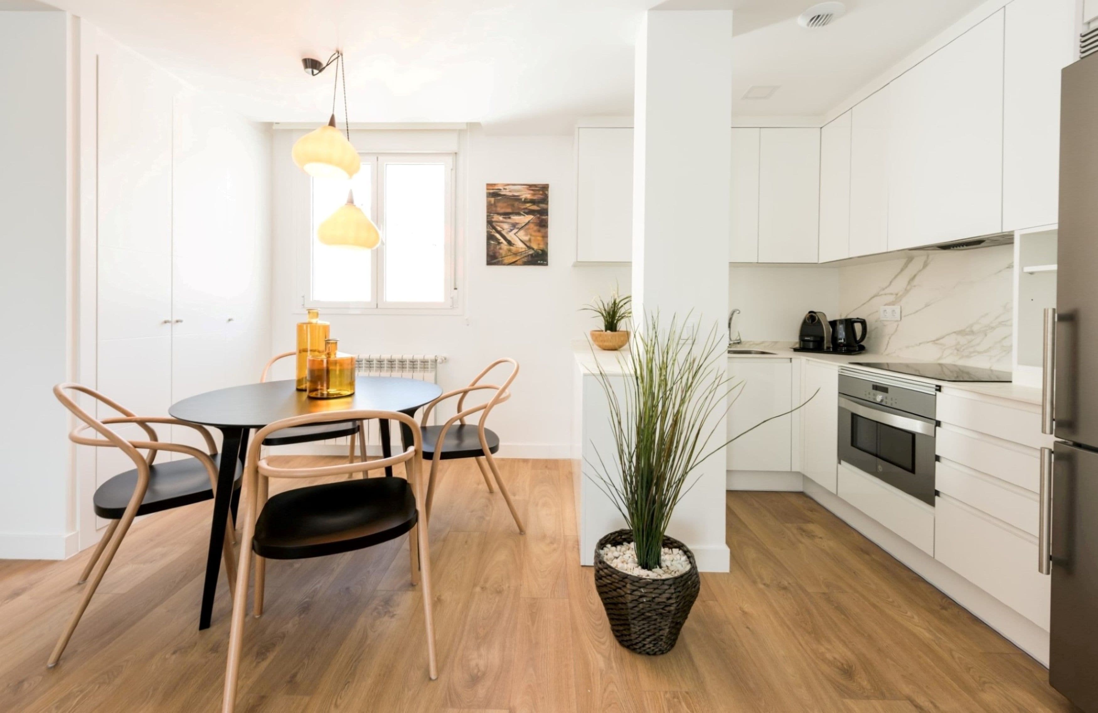 A modern kitchen with white cabinets, a marble countertop, and stainless steel appliances. There is a dining area with a round table and chairs, and a potted plant.