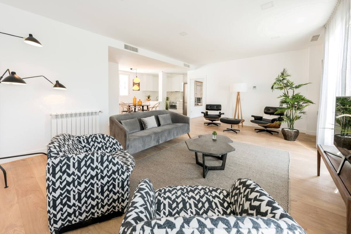 Una sala de estar moderna con un sofá gris, sillones estampados, una mesa de centro y un comedor al fondo.