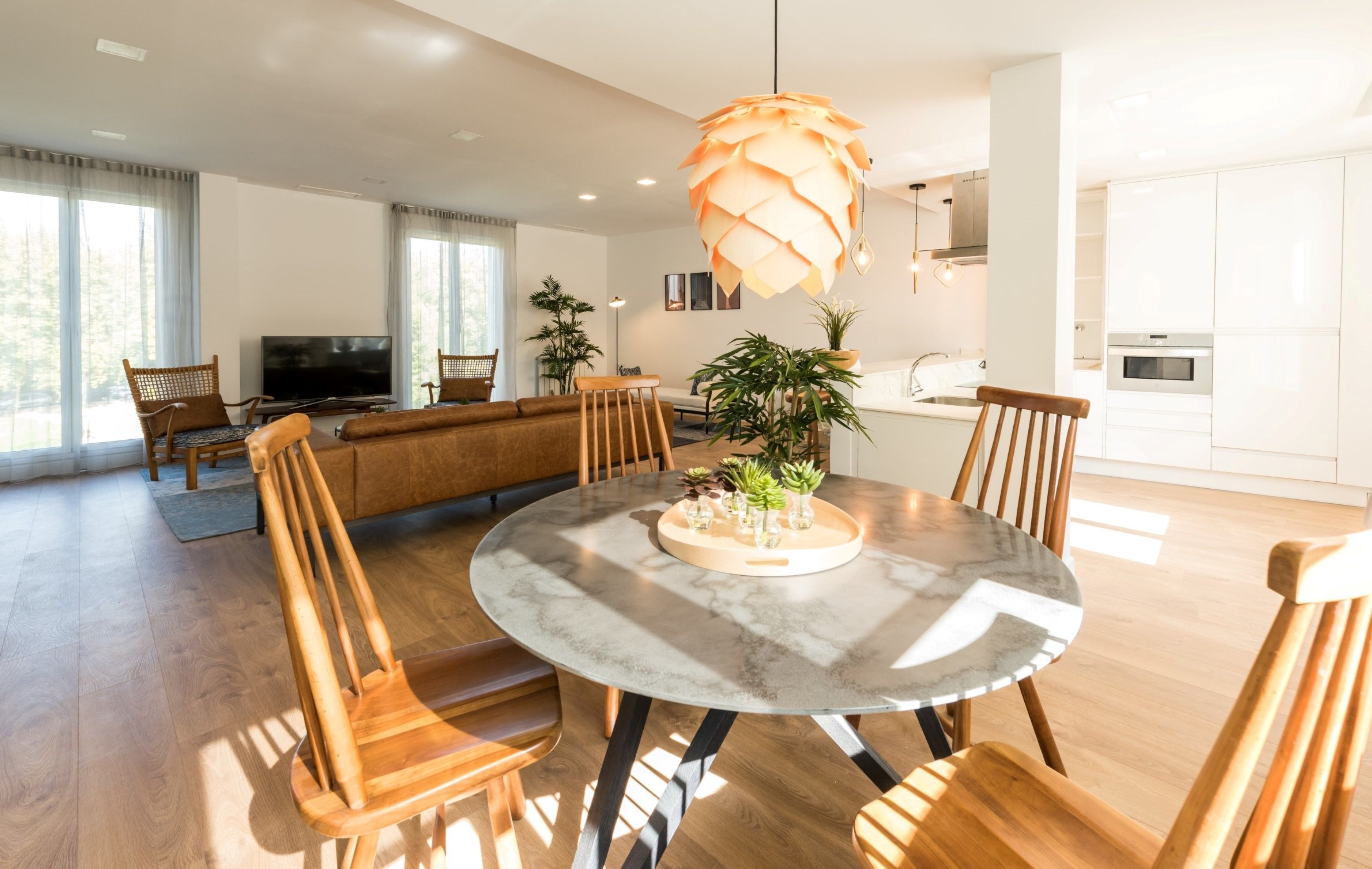 A modern living room with a round dining table, wooden chairs, a TV, and a large window with curtains.