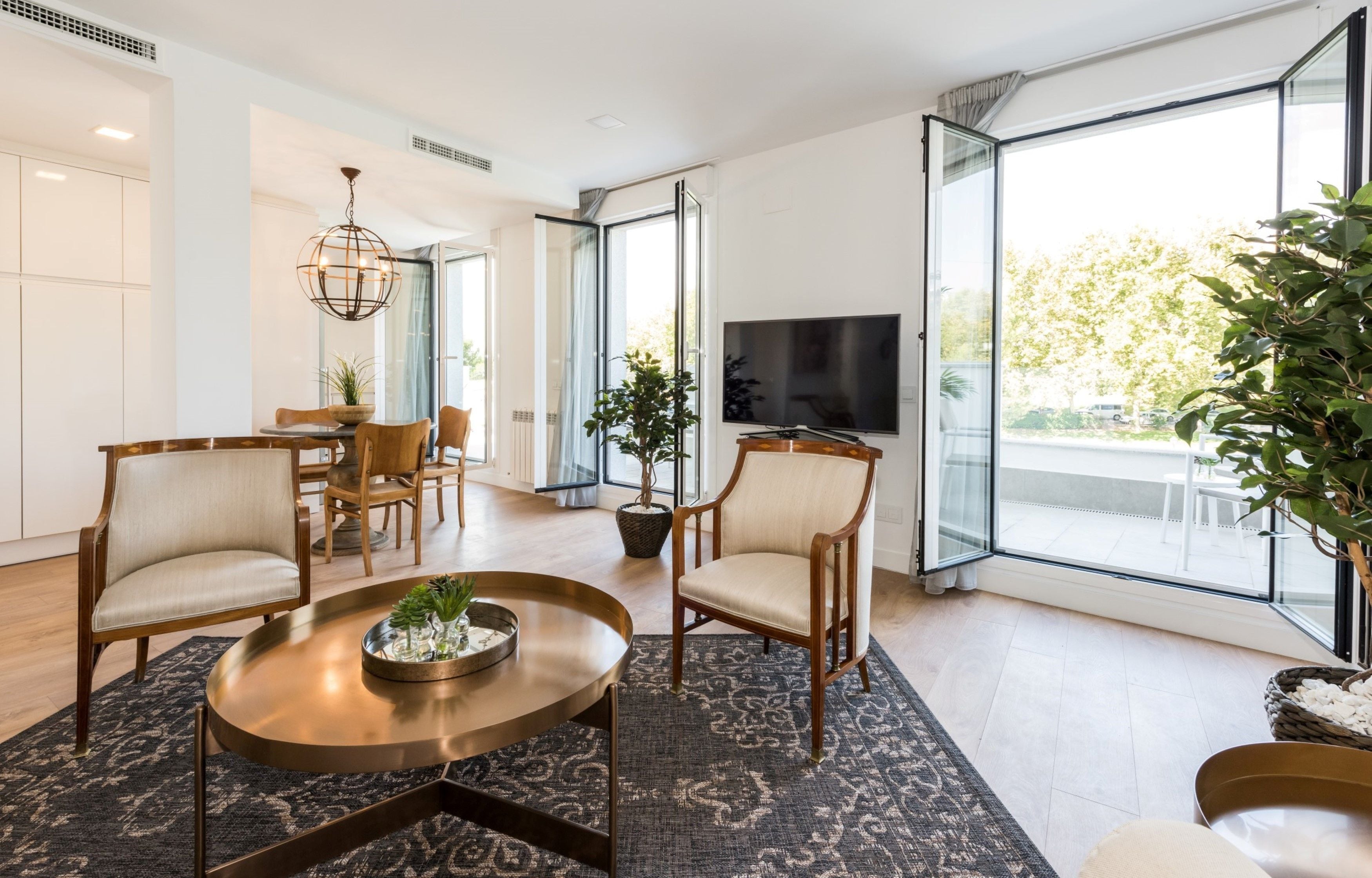 A modern living room with a round coffee table, two armchairs, a dining table with chairs, a TV, and large windows with a view of the outdoors.