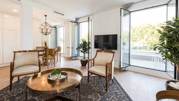 A modern living room with a round coffee table, two armchairs, a dining table with chairs, a TV, and large windows with a view of the outdoors.
