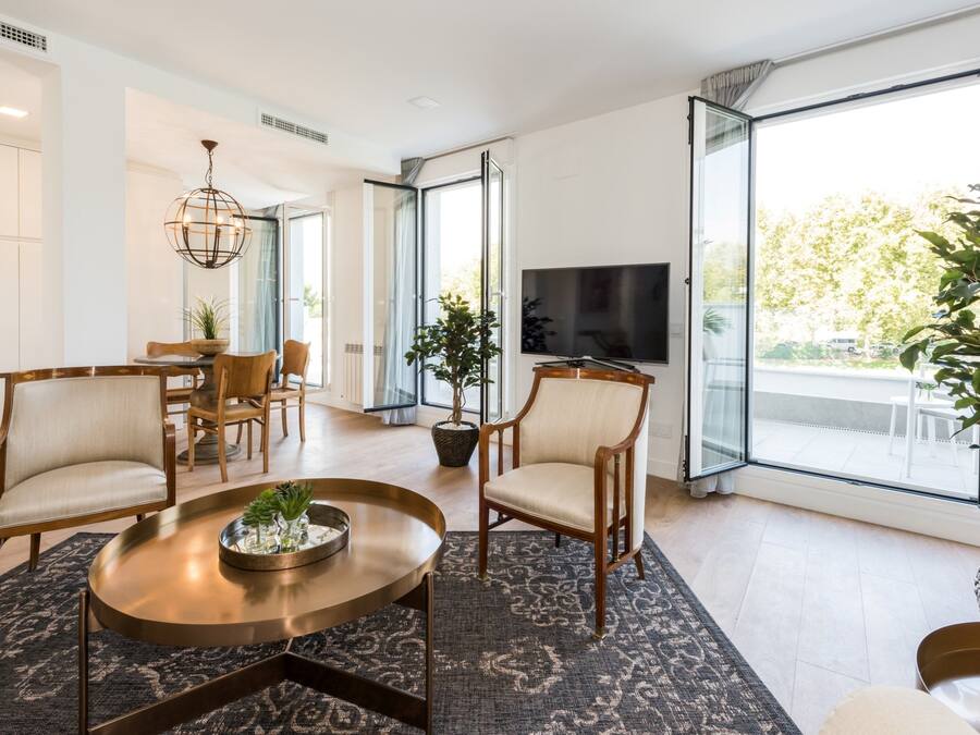 A modern living room with a round coffee table, two armchairs, a dining table with chairs, a TV, and large windows with a view of the outdoors.
