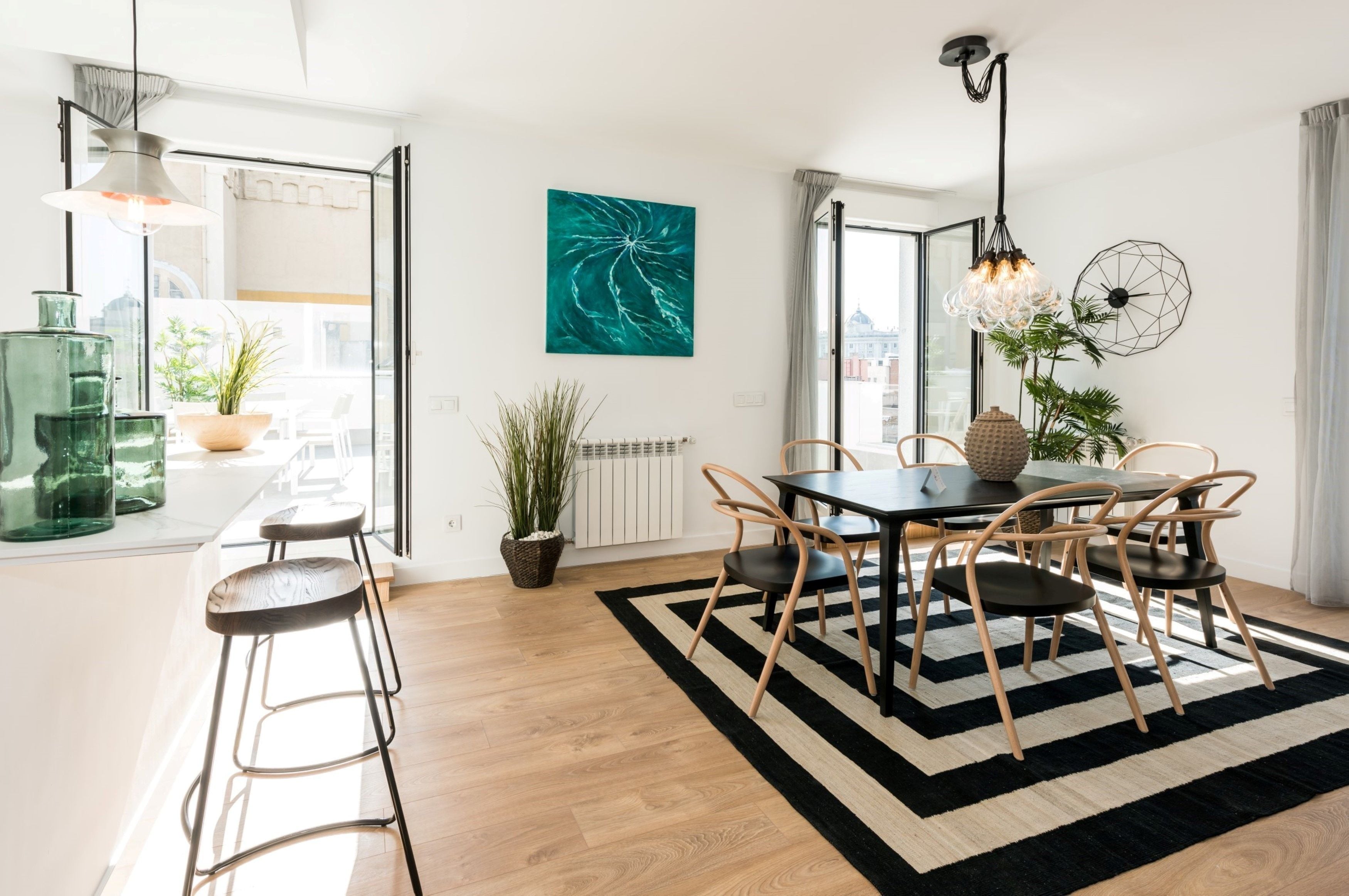 A modern dining area with a black and white striped rug, wooden chairs, and a table. There is a potted plant and a hanging light fixture above the table.