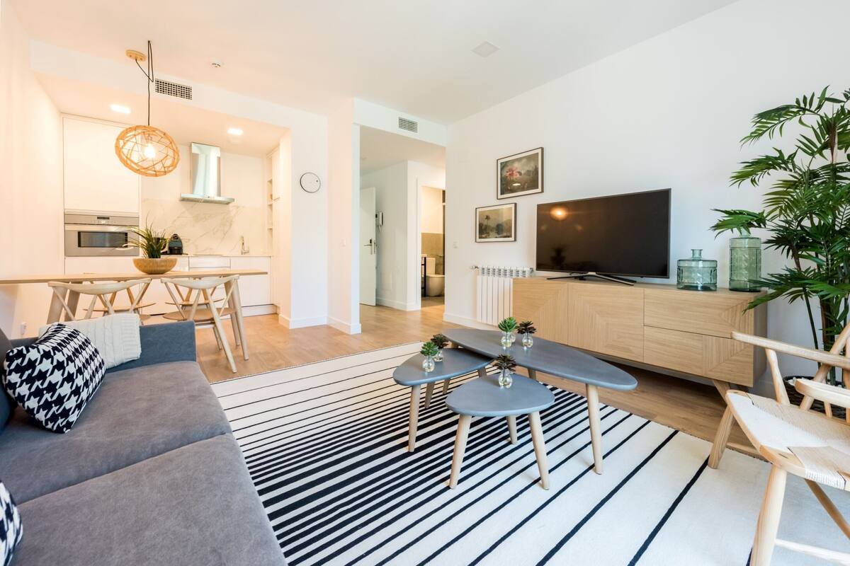 Una sala de estar moderna con un sofá gris, un tapete a rayas negras y blancas, una mesa de comedor de madera con sillas, un televisor y una planta.