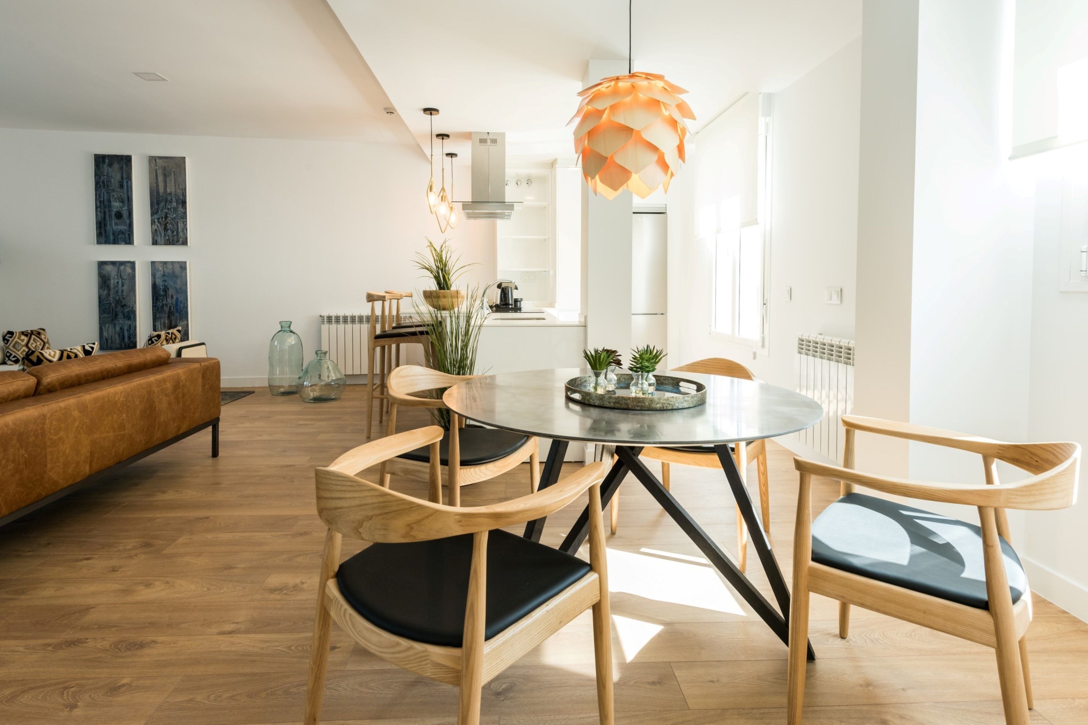 A modern living room with a round dining table, wooden chairs, and a sofa. There are abstract paintings on the wall and a hanging light fixture above the table.