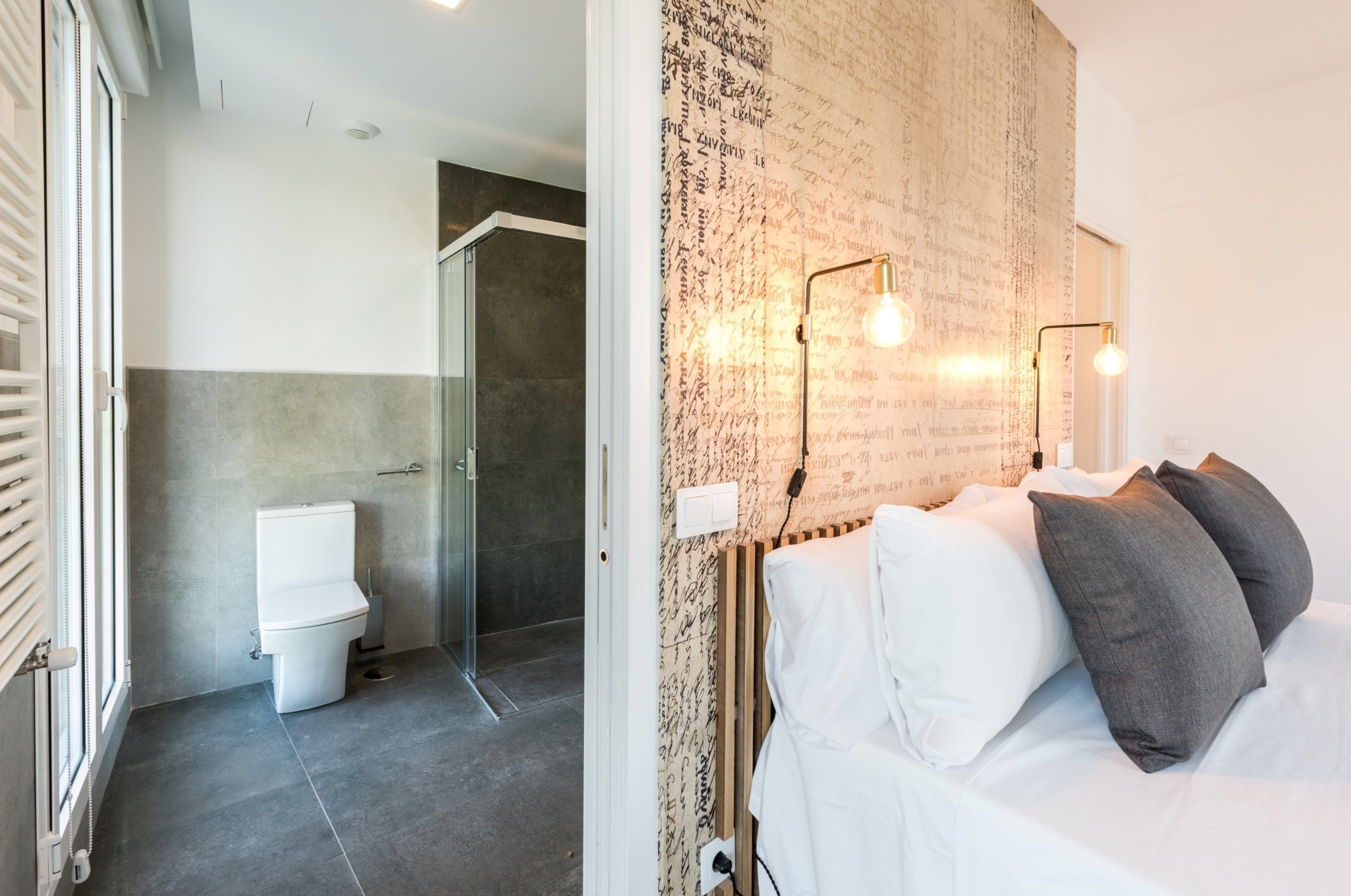 Une salle de bain moderne avec des toilettes, une douche et une porte en verre donnant sur l’espace de la douche. On y trouve un lit avec des oreillers blancs et gris, ainsi qu’un mur recouvert d’un papier peint à motifs.