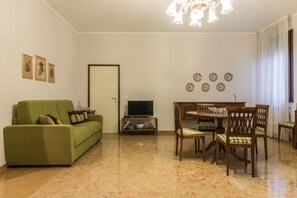 A living room with a green sofa, a dining table with chairs, a TV on a stand, and decorative plates on the wall.