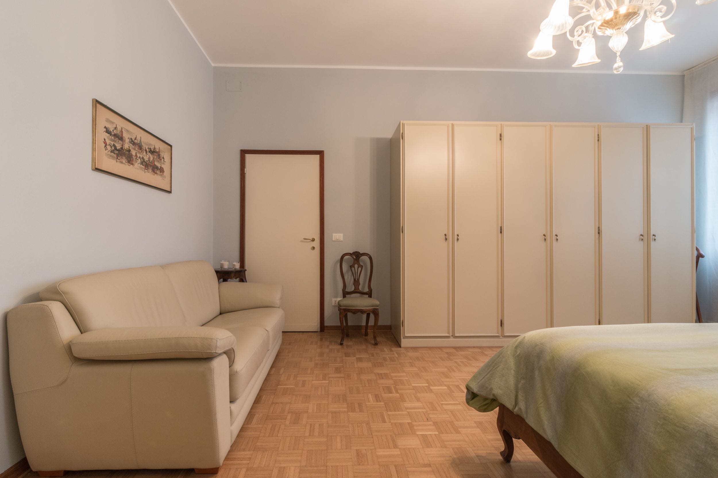 A bedroom with a beige sofa, a wooden chair, a bed with a green bedspread, a wardrobe, and a chandelier.