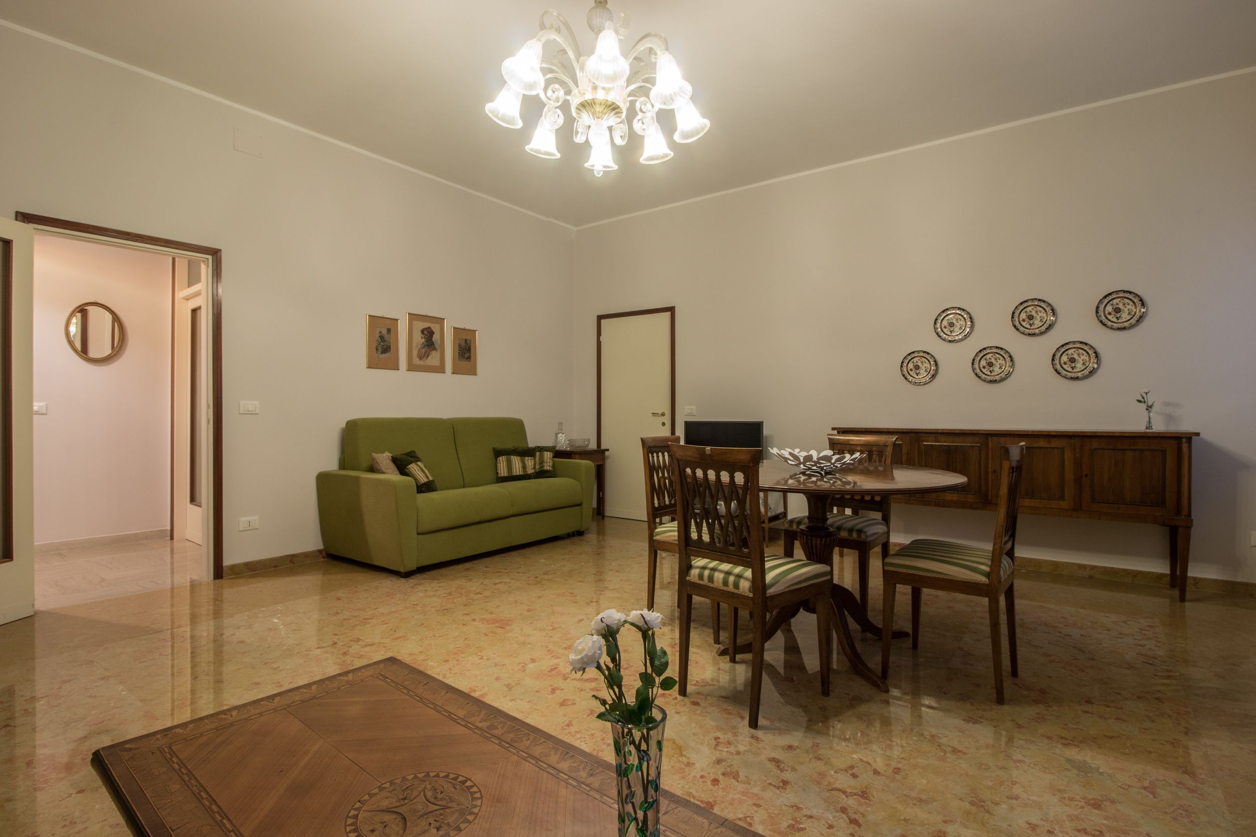 Een woonkamer met een groene bank, een houten eettafel, stoelen, een kroonluchter en decoratieve borden aan de muur.