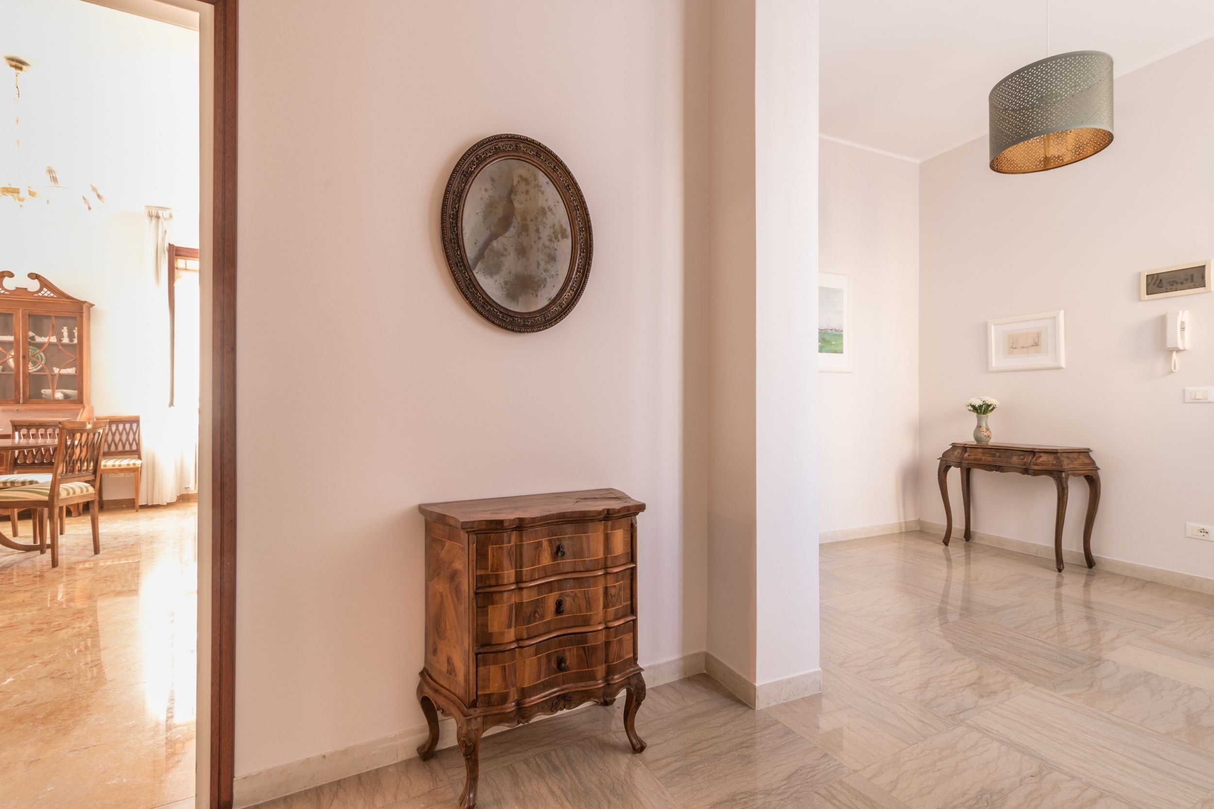 A room with a wooden dresser, a mirror, a table with a vase, and a hanging light fixture.