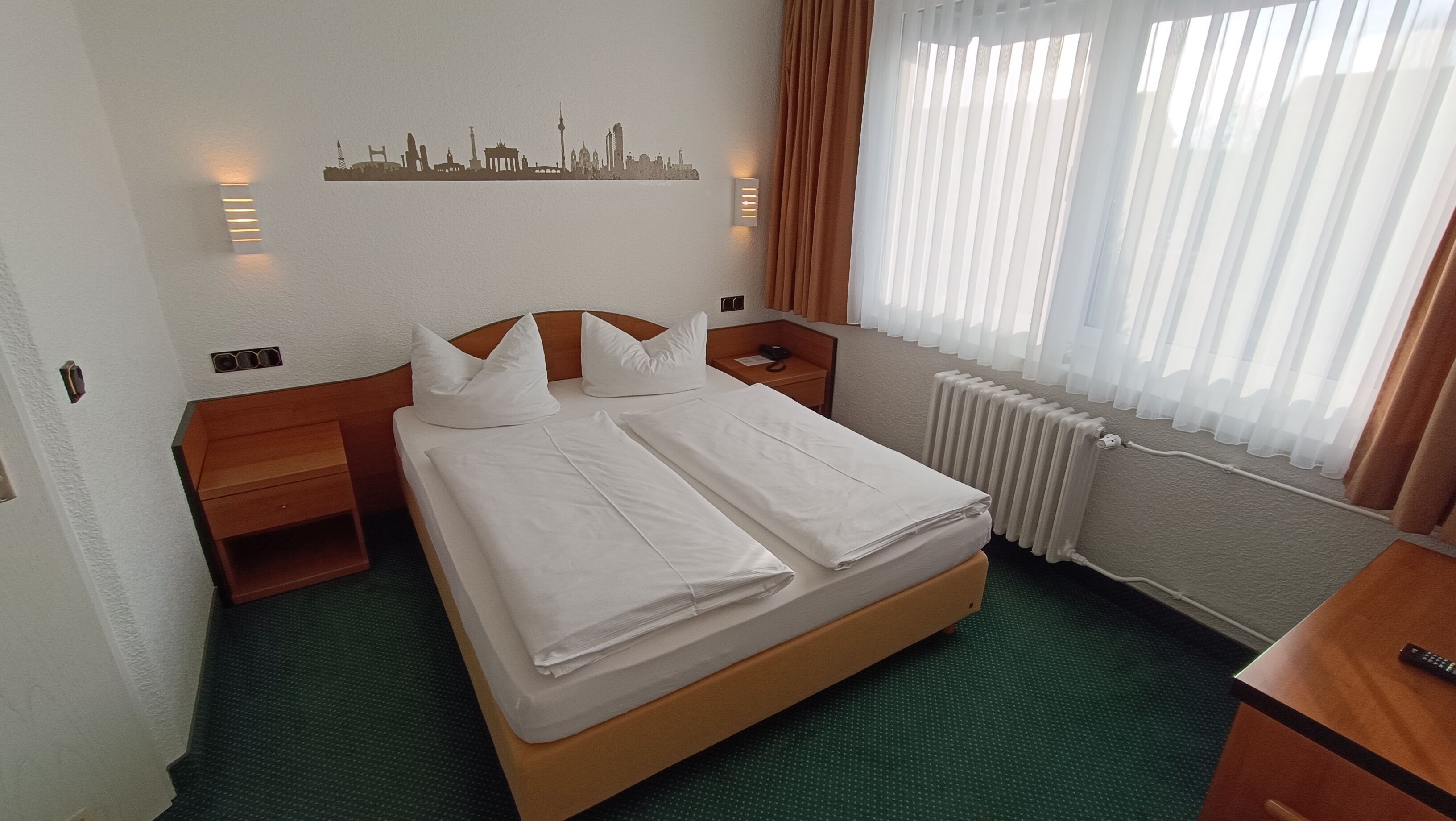 A hotel room with a neatly made bed, a city skyline artwork on the wall, a bedside table with a lamp, and a window with curtains.