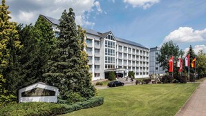 A hotel building with a modern design, a large tree in front, and flags on the right. - LAT Hotel & Apartmenthaus Berlin (Eisenhuettenstadt)
