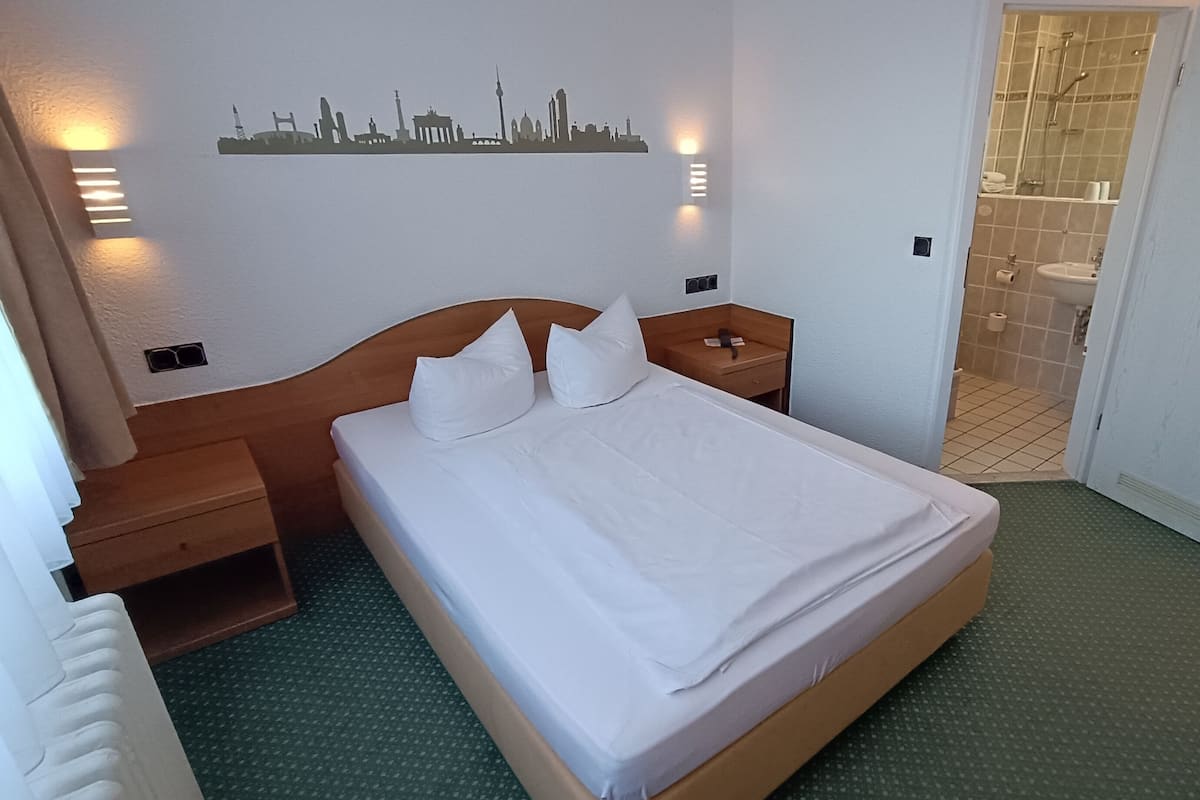 Et hotelværelse med en pænt redt seng, et silhuetbillede af en by skyline på væggen, et badeværelse med flisegulv og en dør, der fører til et andet rum.