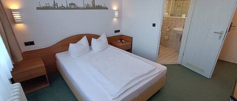 Una habitación de hotel con una cama bien hecha, un perfil silueteado del horizonte urbano en la pared, un baño con piso de baldosas y una puerta que da a otra habitación.
