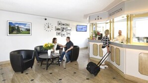 A reception area with a reception desk, a clock, framed pictures on the wall, and a seating area with chairs and a table. - LAT Hotel & Apartmenthaus Berlin (Eisenhuettenstadt)