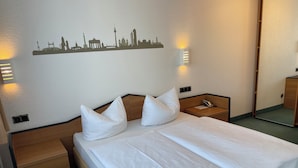 A hotel room with a neatly made bed, a city skyline wall art, bedside lamps, and a small wooden nightstand. - LAT Hotel & Apartmenthaus Berlin (Eisenhuettenstadt)
