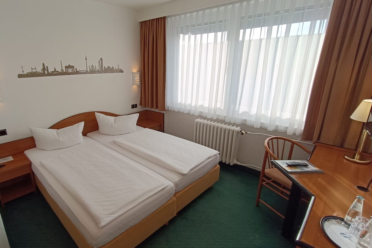 Et hotelværelse med to enkeltsenge, et skrivebord med stol, en radiator og et bybillede på væggen.