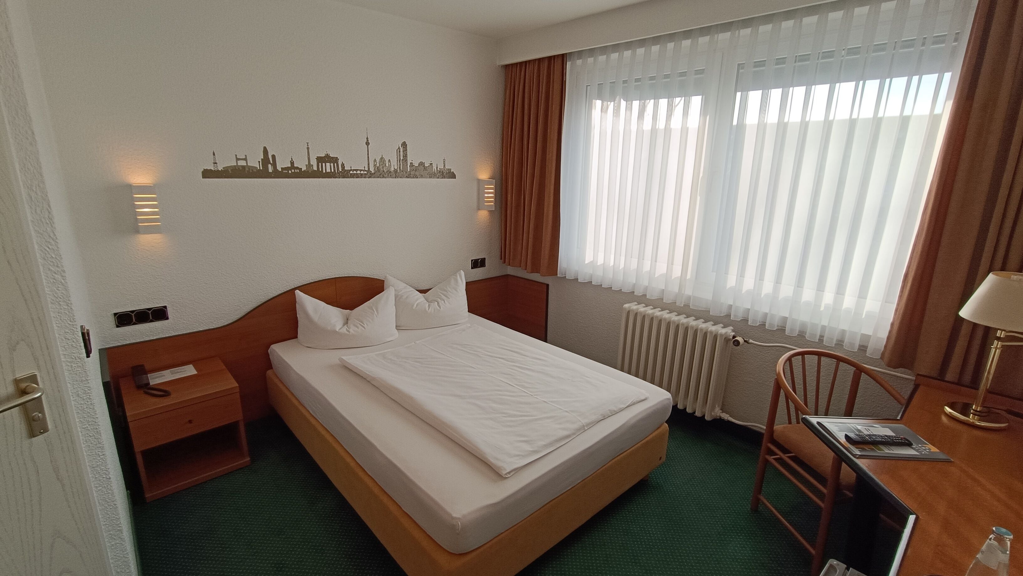 Hotelkamer met een eenpersoonsbed, een bureau met stoel, een klein tafeltje, een radiator en een raam met gordijnen.
