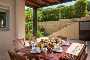 An outdoor dining area with a wooden table set for breakfast, surrounded by chairs, with a view of a landscaped garden and a staircase.