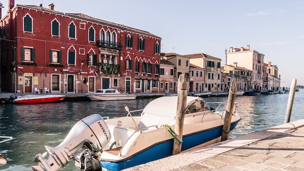 A canal with boats docked, a red building with arched windows, and a row of buildings along the water. - Cà Lessia (Venice)