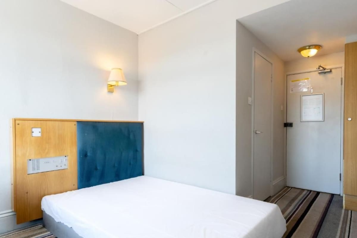 A hotel room with a bed, a wall-mounted light, a door with a sign, and a striped carpet.
