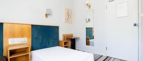 A hotel room with a bed, a wooden wardrobe, a desk, a mirror, and a wall-mounted light fixture.