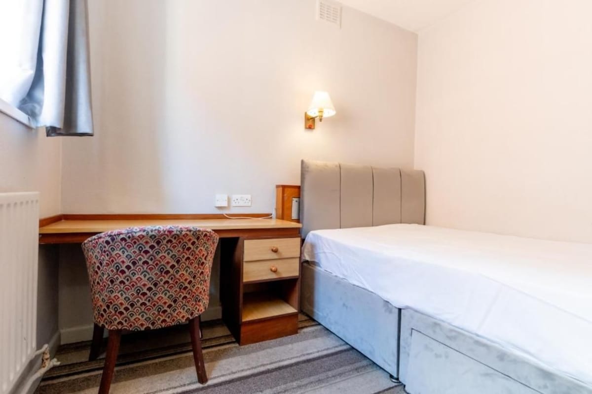 A small, clean room with a bed, a desk, and a chair. There is a wall-mounted light above the desk.