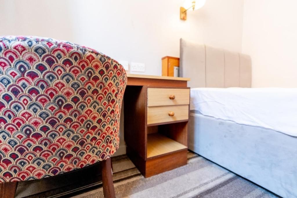 A hotel room with a bed, a wooden nightstand with drawers, and a patterned chair.