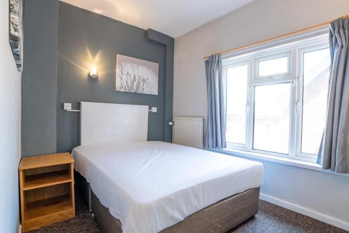 A bedroom with a bed, a nightstand, a radiator, and a window with curtains.