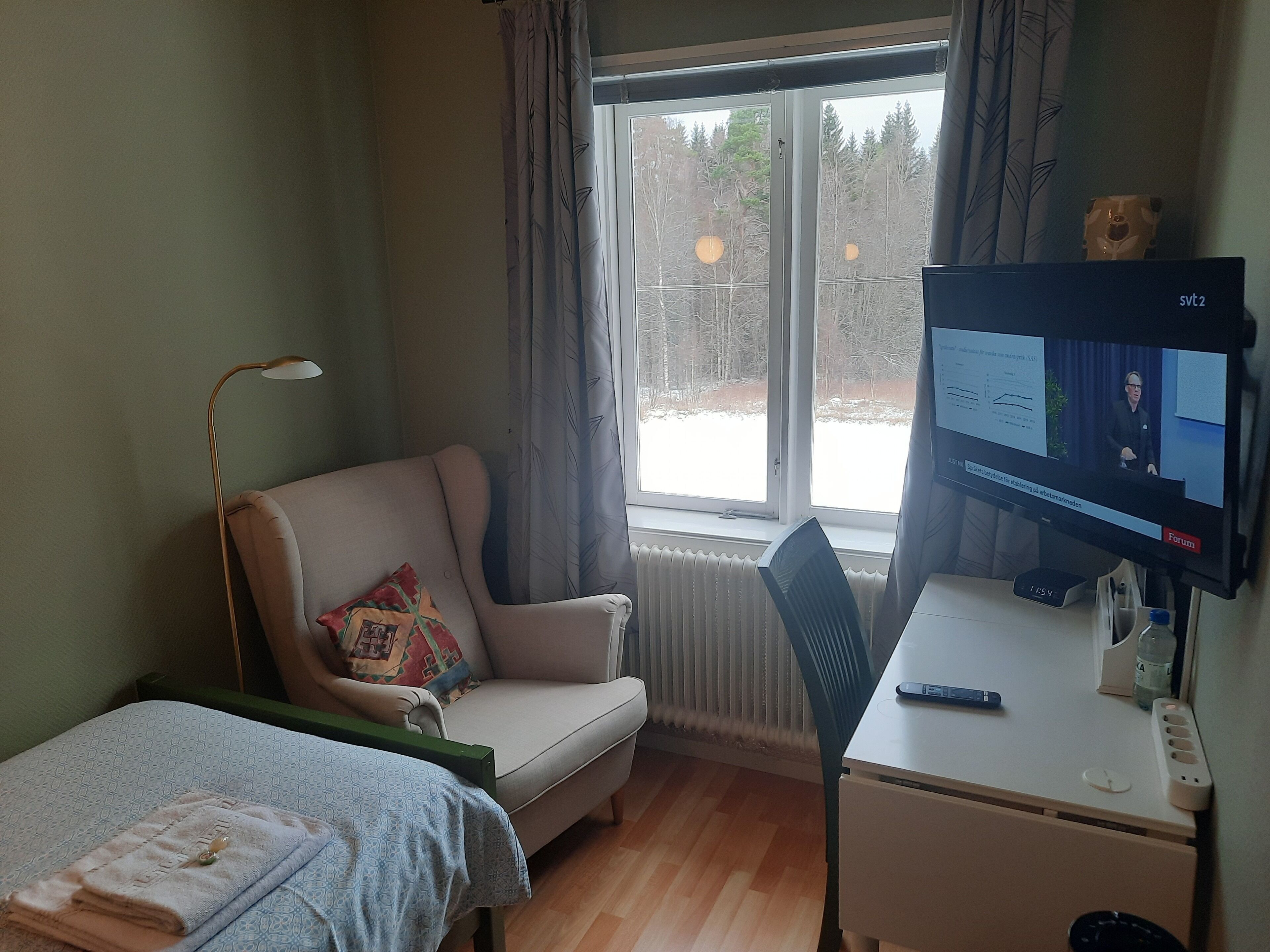 A room with a bed, a chair, a desk with a computer, and a window with curtains.