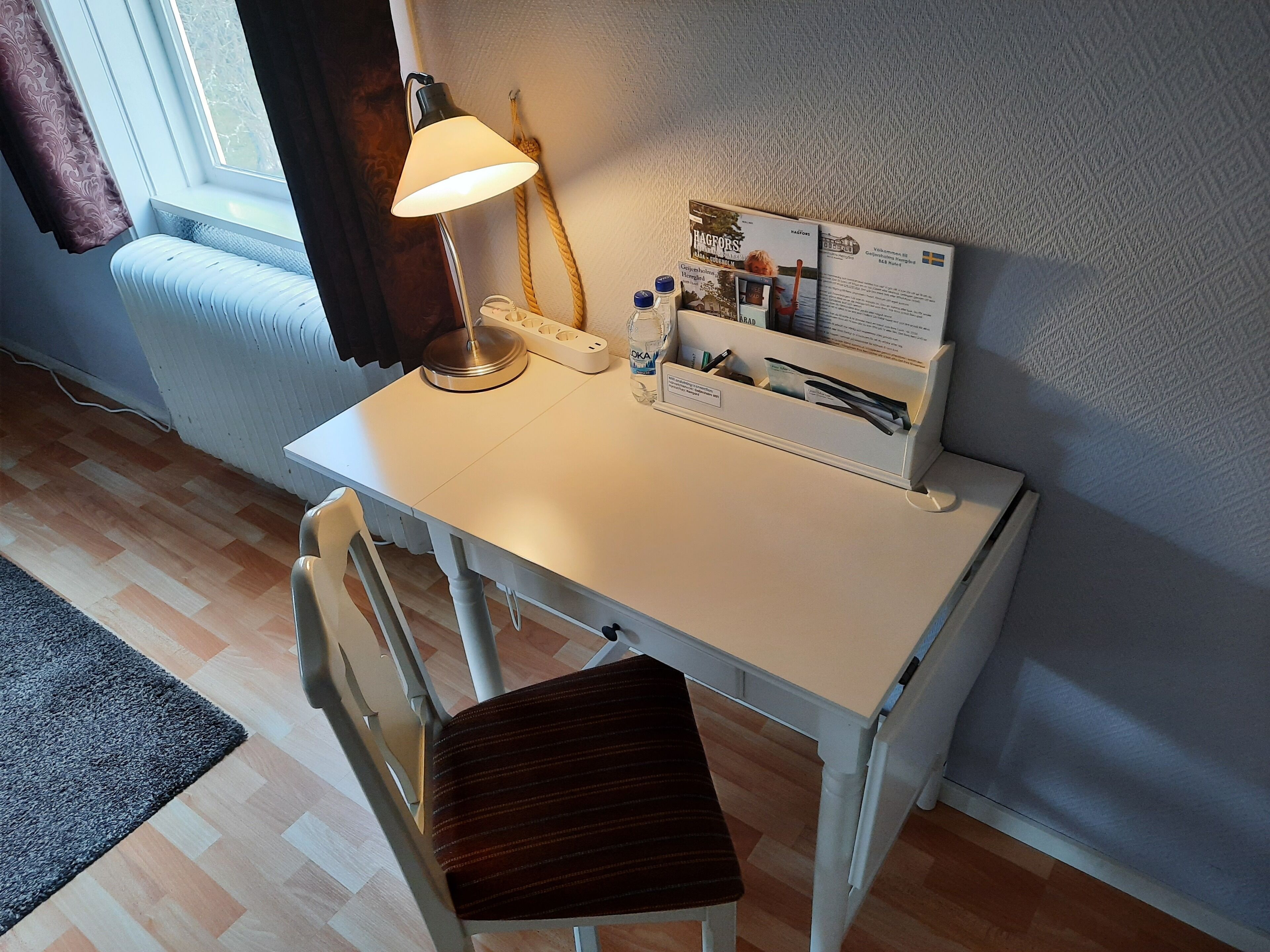 Een klein wit bureau met een lamp, een stoel en een brochure op tafel.