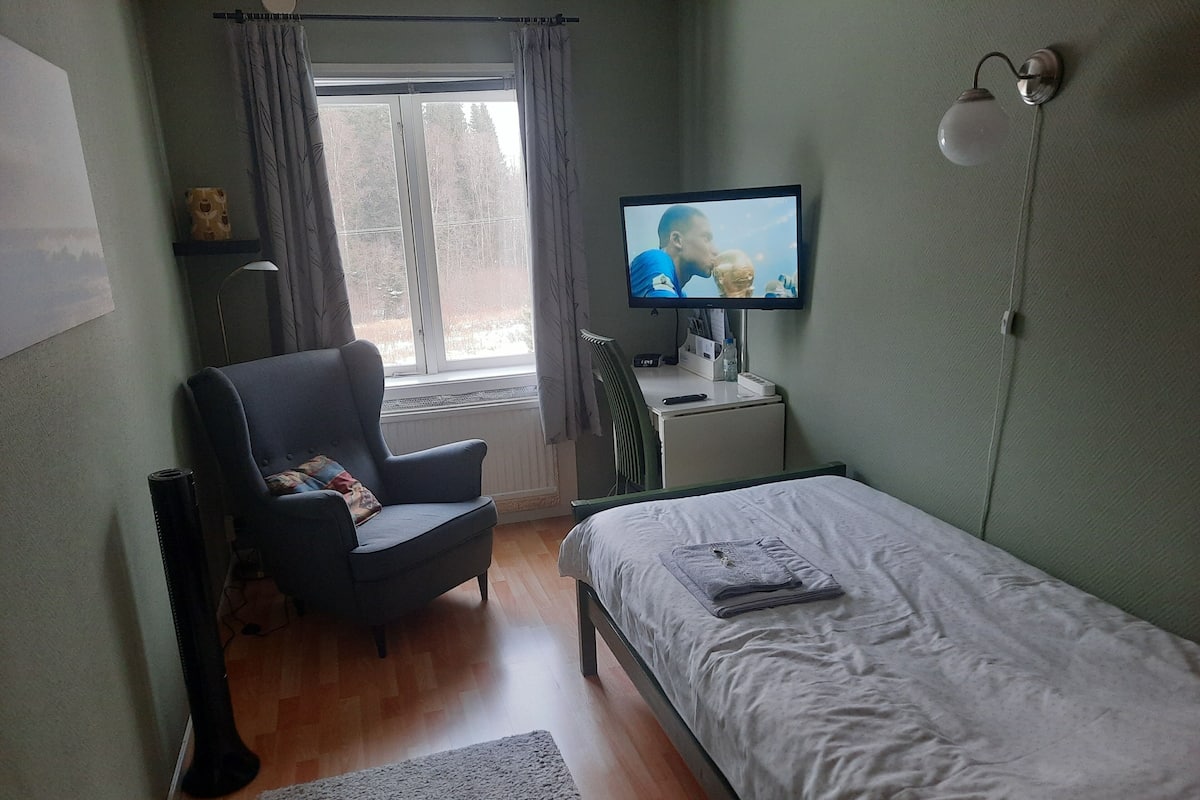 Een slaapkamer met een bed, een stoel, een bureau met een computer, een raam met gordijnen, en een televisie aan de muur.